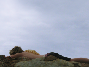 A light-skinned body lying on their front on rocks by the beach, with a fish fin protruding from their back.